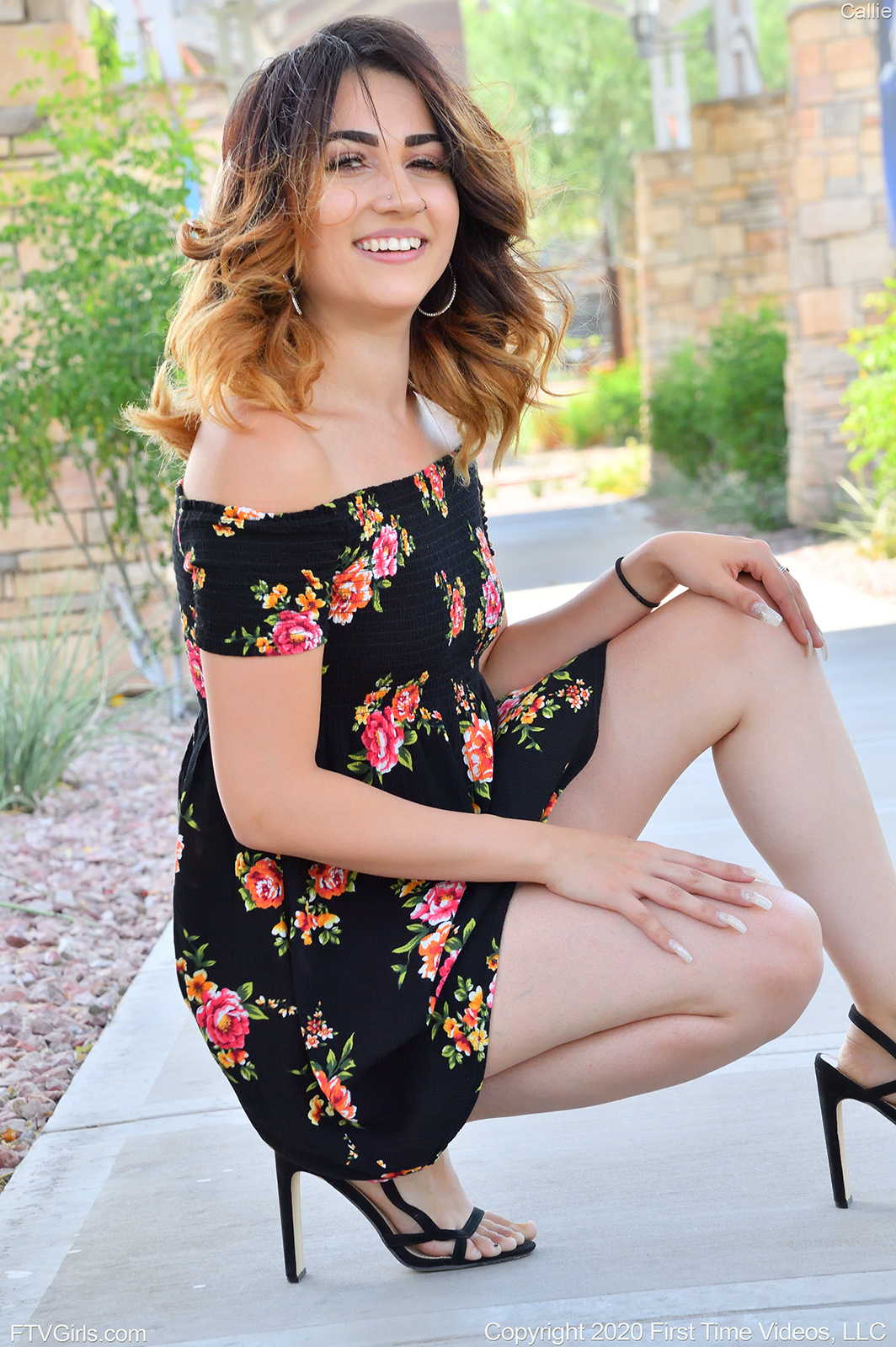 teen pornstar Callie FTV is non nude wearing a black floral dress and high heels while in squat position on a concrete floor outdoors and smiling at the camera in Girl Next Door Style
