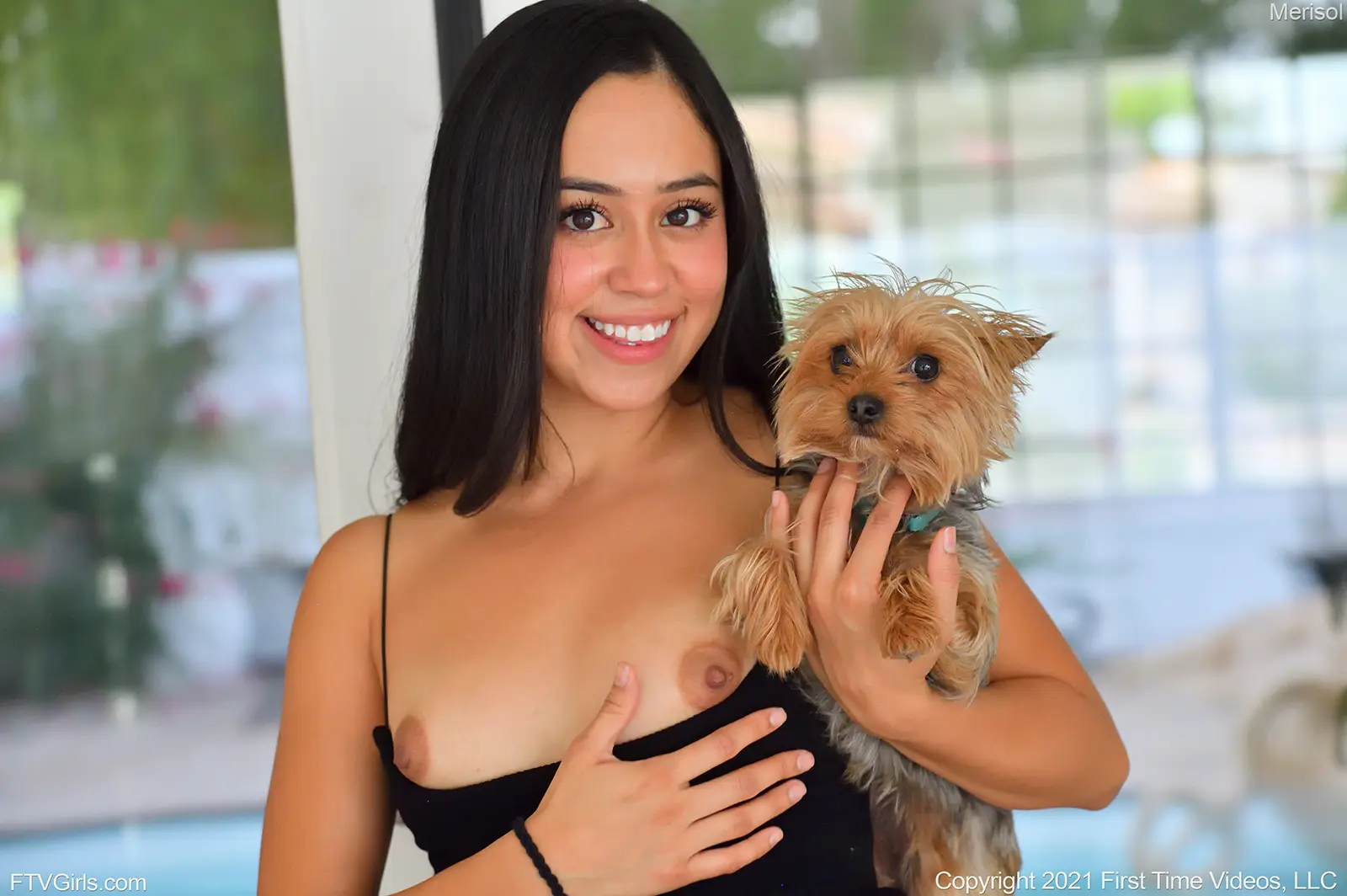 Merisol FTV is holding a beautiful dog as she hows her tits while standing near a glass door near the pool