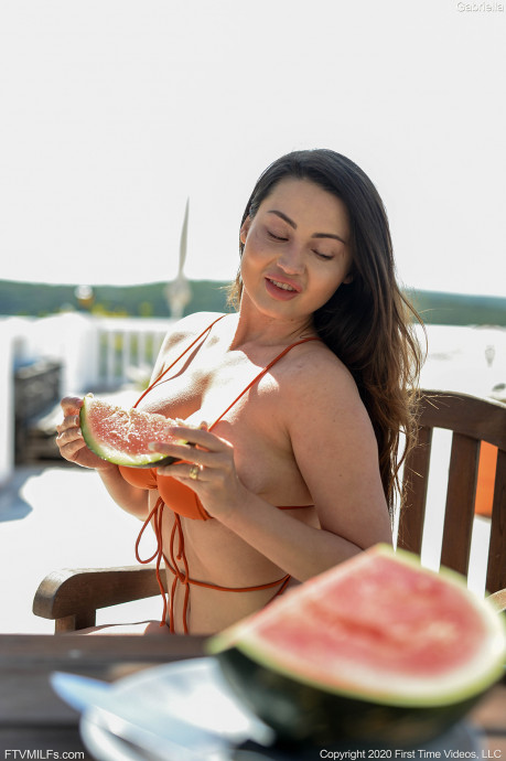 milf pornstar Gabriella FTV is holding a watermelon while sitting on a wooden chair wearing a bikini in Melon Juice